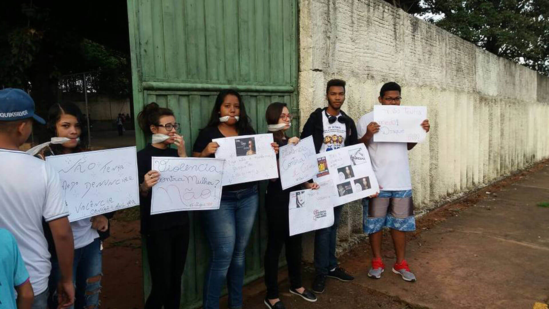 Imagem de alunos protestando com mordaças na boca e cartazes nas mãos em frente ao portão de entrada da escola